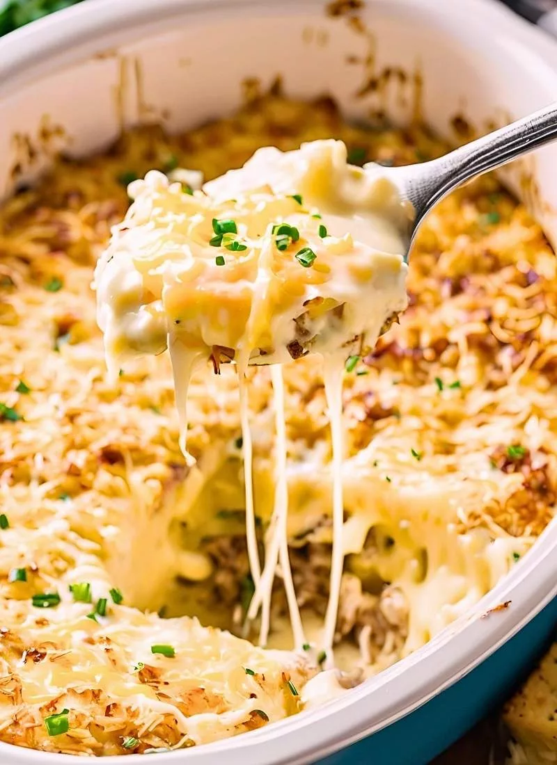 A heaping spoonful of slow cooker hashbrown casserole being lifted, revealing gooey layers of melted cheese, tender shredded potatoes, and creamy sauce. The cheese stretches slightly, emphasizing