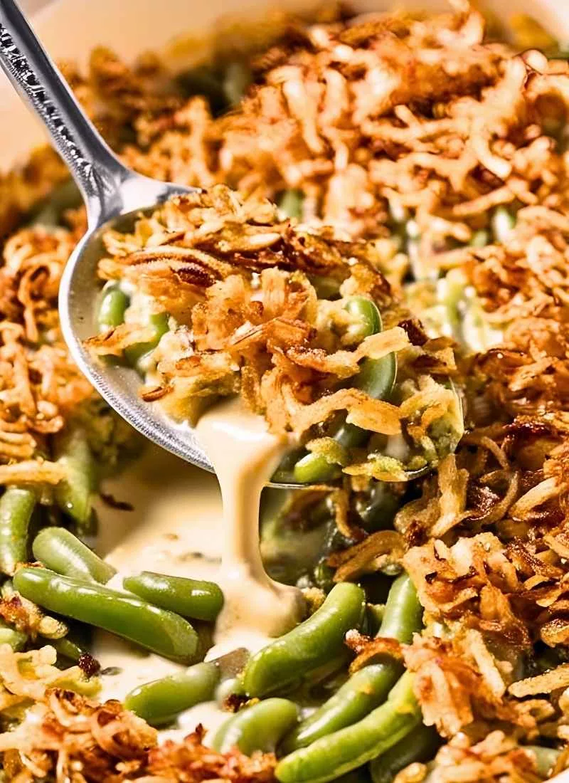 A spoon lifting a heaping scoop of Campbell’s Green Bean Casserole, revealing the rich, creamy sauce coating tender green beans. The crispy fried onions sit on top, slightly crumbling as the spoon