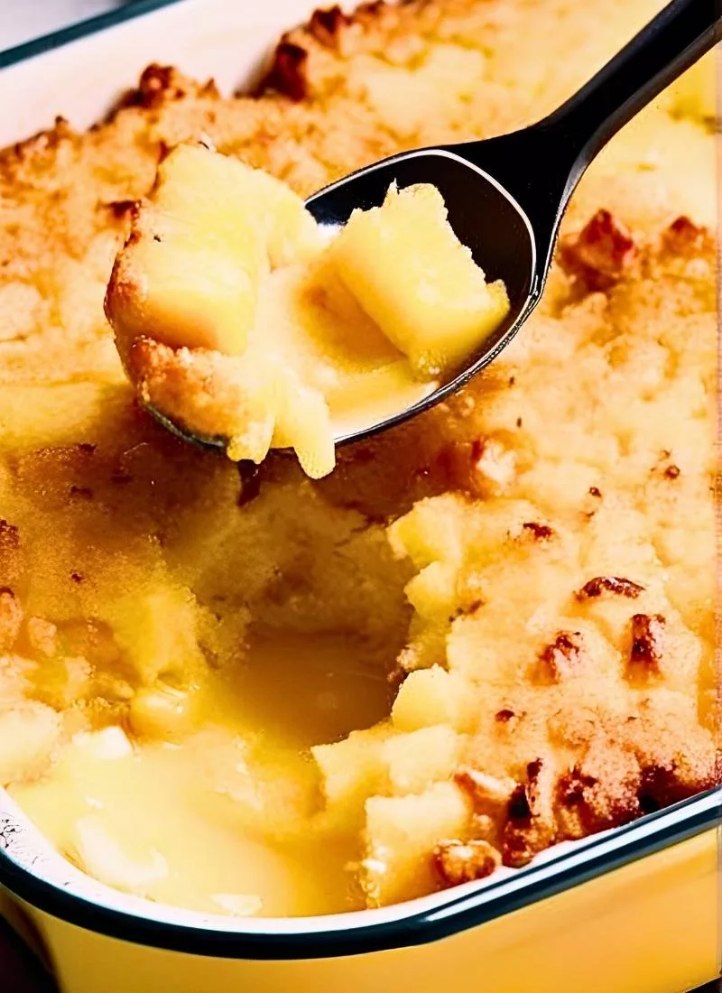 Spoon lifting a scoop of Pineapple Casserole from the baking dish – Showcases the soft custard like filling with juicy chunks of pineapple and a crispy buttery topping ()