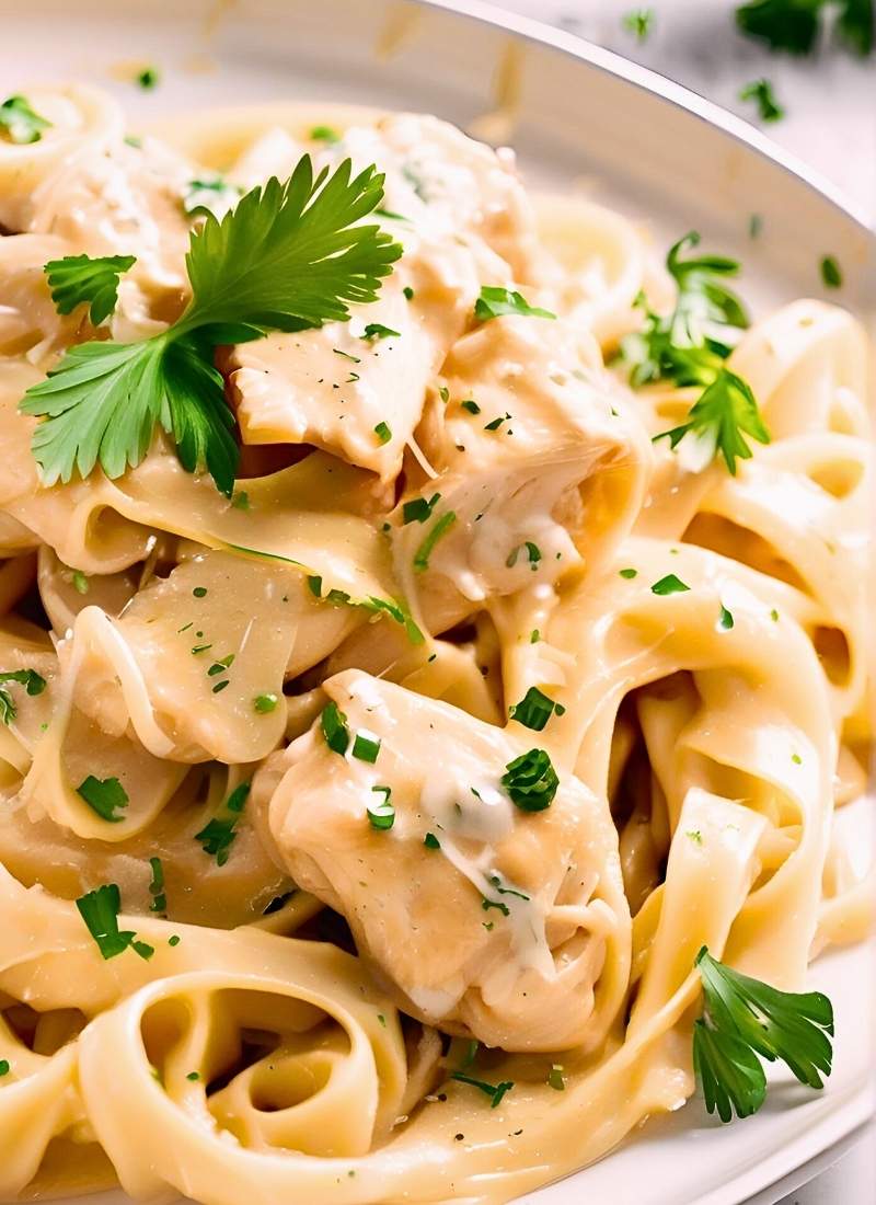 Twirled fettuccine in a glossy, creamy garlic sauce, golden chicken chunks glistening, flecked with fresh parsley, and a sprinkle of melting parmesan, set on a rustic white plate with soft light. (1)
