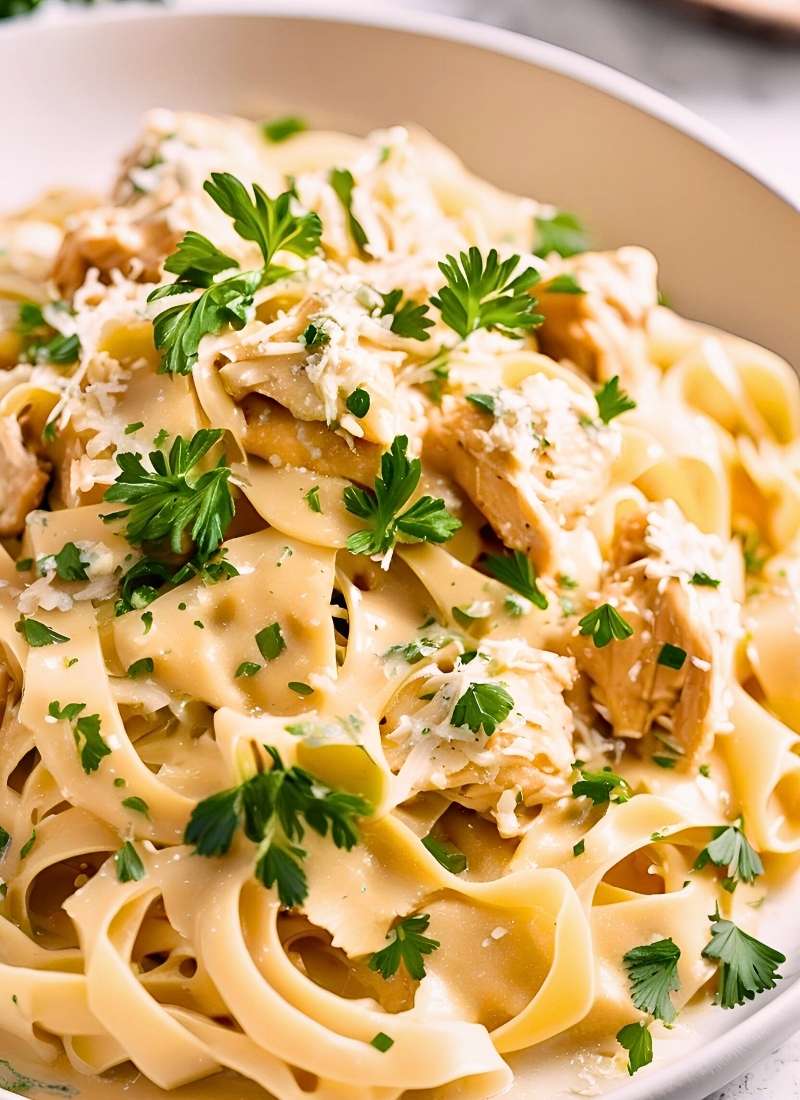 Twirled fettuccine in a glossy, creamy garlic sauce, golden chicken chunks glistening, flecked with fresh parsley, and a sprinkle of melting parmesan, set on a rustic white plate with soft light.